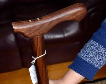 May include: A wooden walking stick with a brown and black wood grain pattern. The handle is curved and has a thin strip of orange wood inlay. A white tag is attached to the stick with the text "$210.00" and "Walnut" written on it.