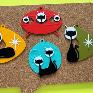 May include: Five retro-style cat ornaments with a mid-century modern design. The ornaments are made of plastic and feature black cats with white faces and antennas. The ornaments are in various shapes, including a flying saucer, a moon, and a boomerang.