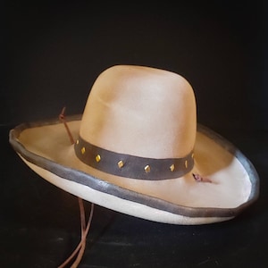 Könnte beinhalten: Ein breitkrempiger, cremefarbener Filzhut im Cowboy-Stil mit einem braunen Lederband und goldenen Nieten. Der Hut hat eine leicht spitze Krone und eine breite, flache Krempe.