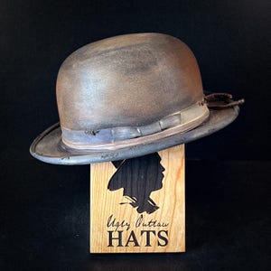 May include: A brown bowler hat with a distressed finish and a black band. The hat is displayed on a wooden stand with the text "Ugly Outlaw HATS" engraved on it.