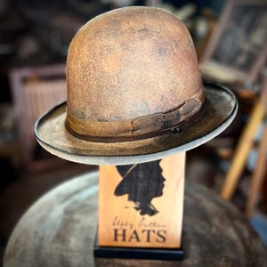 May include: A vintage-style brown bowler hat with a dark brown ribbon band. The hat is displayed on a wooden stand with a silhouette of a person wearing a hat and the text "Wisty Button Hats". The hat has a slightly distressed appearance.