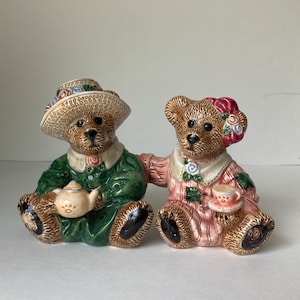 May include: Two ceramic teddy bear figurines. One bear wears a green dress and hat, holding a small teapot. The other bear wears a pink dress and a floral headpiece, holding a teacup. Both bears have brown fur and black paws.