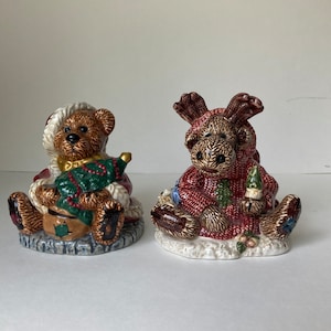 May include: Two ceramic Christmas figurines, a brown bear wearing a Santa hat and holding a small Christmas tree, and a brown moose wearing a red sweater and holding a small Christmas tree.