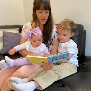 May include: A woman reads a book titled "Norman and the Temple" to two children, one sitting on her lap and the other sitting beside her on a black leather couch.