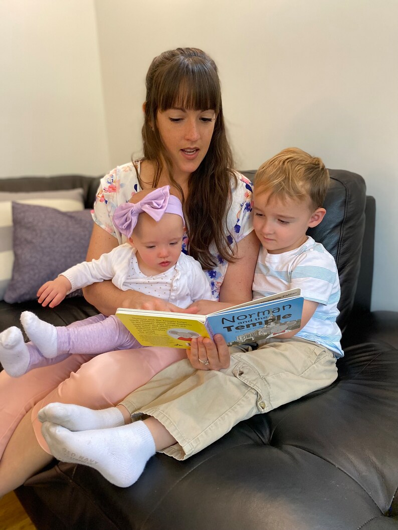 May include: A woman reads a book titled "Norman and the Temple" to two children, one sitting on her lap and the other sitting beside her on a black leather couch.