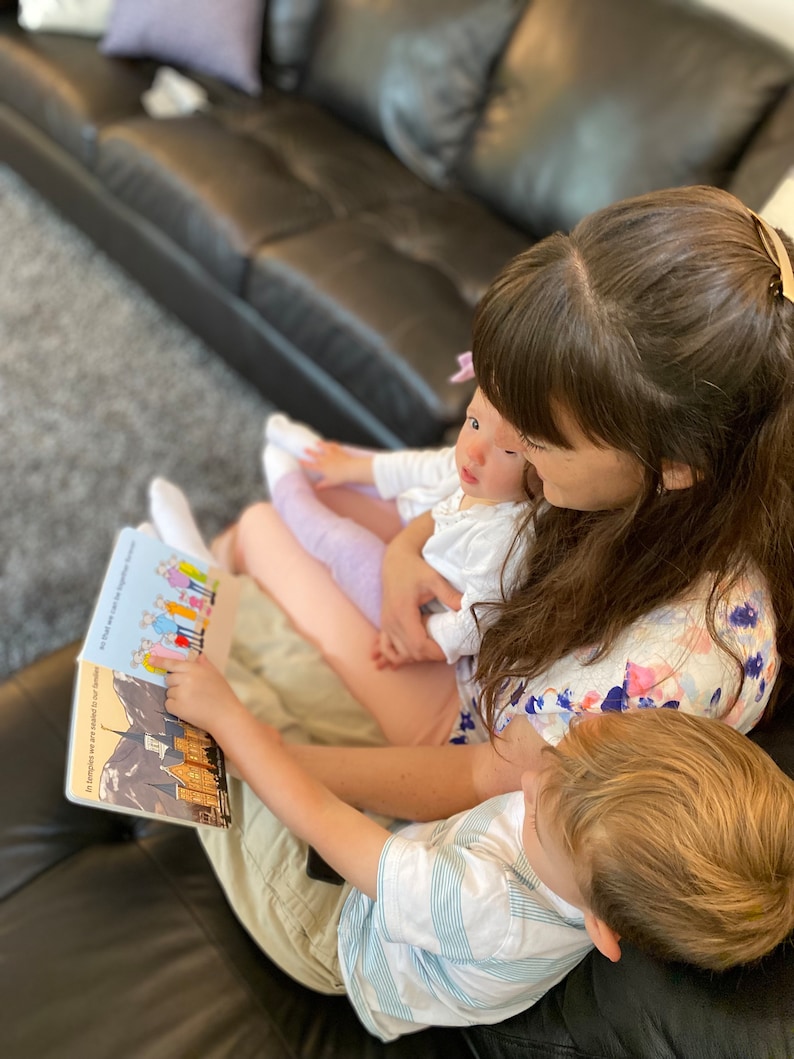 May include: A woman sits on a couch with two children, reading a book titled "In Chicago, we are treated to number". The book features illustrations of people and buildings. The woman is wearing a floral shirt and the boy is wearing a striped shirt.