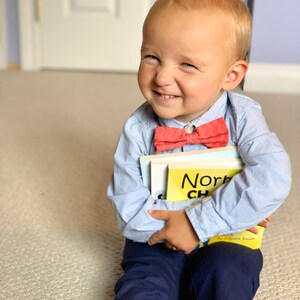 May include: A young child wearing a light blue striped button-down shirt, navy blue pants, and a coral bow tie. The child is holding books, one of which reads "Norbert". The child is winking and smiling.