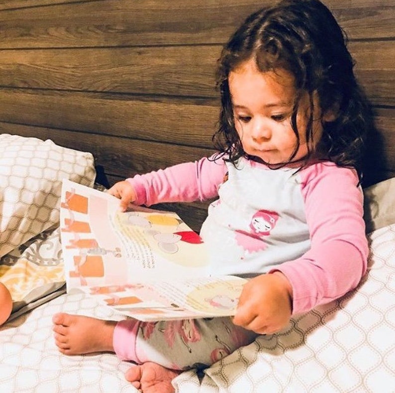May include: A young child in pink pajamas sits on a bed and reads a book with colorful illustrations. The book is open to a page with a picture of a pink flamingo.