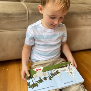 May include: A young boy sits on a wooden floor, reading a children's book about feeling happy and peaceful. The book features a cartoon mouse with a red bow tie and a white temple building.