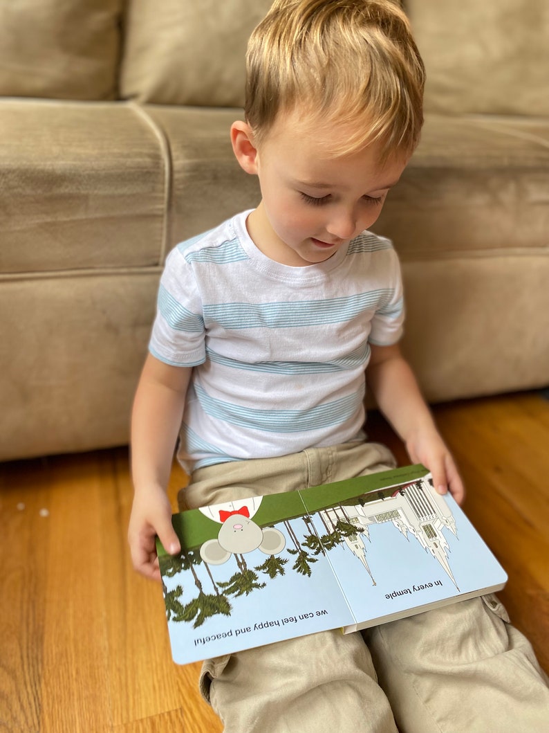 May include: A young boy sits on a wooden floor, reading a children's book about feeling happy and peaceful. The book features a cartoon mouse with a red bow tie and a white temple building.
