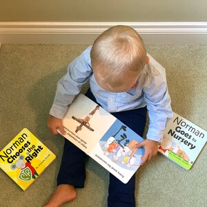 May include: A child in a light blue shirt and navy pants is surrounded by three children's books. The books are titled "Norman Chooses the Right" and "Norman Goes to Nursery". The open book shows illustrations and text.