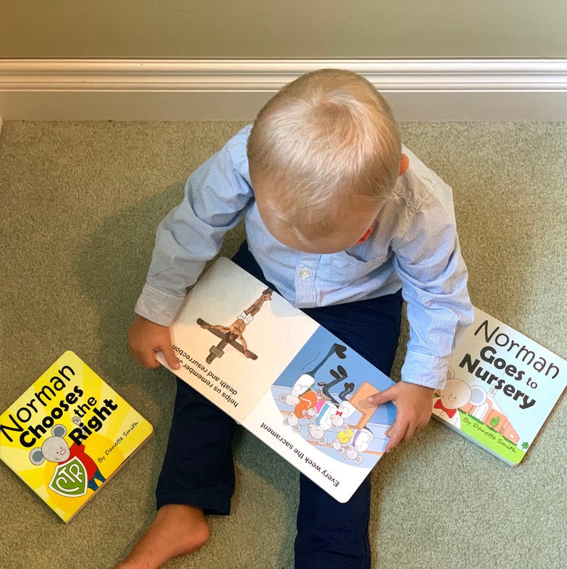 May include: A child in a light blue shirt and navy pants is surrounded by three children's books. The books are titled "Norman Chooses the Right" and "Norman Goes to Nursery". The open book shows illustrations and text.