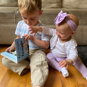 May include: Two children, a boy and a girl, are sitting on a wooden floor. The boy is reading a book titled "Norman and the Temple" to the girl. The girl is wearing a white shirt with gold polka dots, purple pants, and a purple bow headband. The boy is wearing a blue and white striped shirt, khaki pants, and white socks.