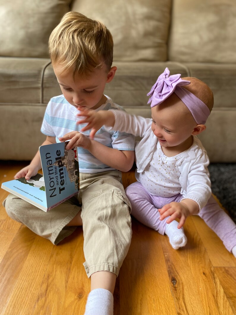 May include: Two children, a boy and a girl, are sitting on a wooden floor. The boy is reading a book titled "Norman and the Temple" to the girl. The girl is wearing a white shirt with gold polka dots, purple pants, and a purple bow headband. The boy is wearing a blue and white striped shirt, khaki pants, and white socks.