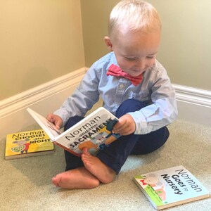 May include: A young child sits on a carpet, reading a book titled "Norman and the Sacrament." Two other books, "Norman Chooses the Right" and "Norman Goes to Nursery," are nearby. The child wears a light blue shirt, navy pants, and a red bow tie.