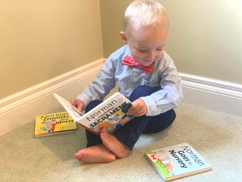 May include: A young child sits on a carpet, reading a book titled "Norman and the Sacrament." Two other books, "Norman Chooses the Right" and "Norman Goes to Nursery," are nearby. The child wears a light blue shirt, navy pants, and a red bow tie.