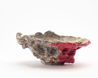 Puede incluir: Un pequeño cuenco de cerámica de forma irregular con una superficie rugosa y texturizada. El cuenco está pintado con un esmalte rojo brillante en un lado, y el resto del cuenco es gris y marrón moteado.