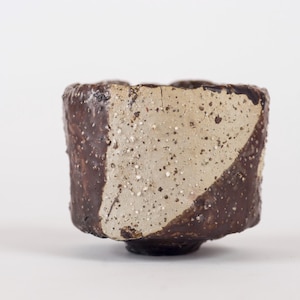 Peut inclure: Une tasse en céramique carrée faite à la main avec un design unique. La tasse présente une section diagonale d'argile claire mouchetée contre un extérieur brun foncé texturé. La base est un petit pied arrondi.