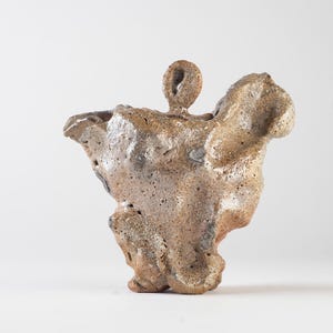 Op de afbeelding: Een handgemaakte, abstracte keramische sculptuur met een ruw, getextureerd oppervlak. De sculptuur is voornamelijk bruin met hints van grijs en zwart, en heeft een organische, asymmetrische vorm. Het stuk heeft een kleine, cirkelvormige opening aan de bovenkant.