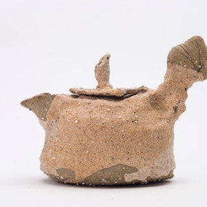 Peut inclure: Une petite théière en céramique de couleur sable avec une surface rugueuse et texturée. La théière a une forme unique et irrégulière avec un bec verseur et un couvercle.