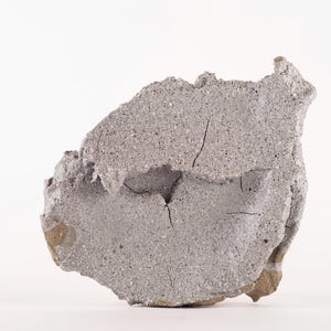 Peut inclure: Un morceau de béton gris de forme irrégulière avec des fissures visibles et une texture rugueuse. L'objet est posé sur un fond blanc, soulignant sa forme et ses détails. Le béton présente des tons gris clairs et foncés.