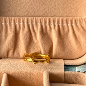 May include: A gold ring with a delicate, twig-like design. The ring is displayed in a beige velvet jewellery box.