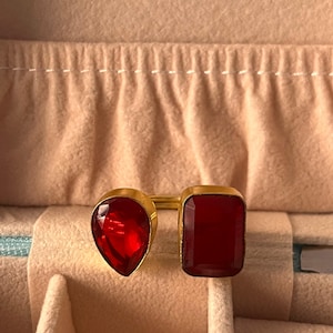 May include: Two gold rings with large red gemstones. One ring features a teardrop-shaped stone, and the other has a rectangular stone. The rings are displayed inside a beige jewelry box.