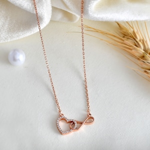 Könnte beinhalten: Halskette aus Roségold mit einem Herz- und Unendlichkeitssymbol-Anhänger. Das Herz ist mit kleinen, klaren Kristallen besetzt. Die zarte Kette hängt vor einem weißen Hintergrund, mit einer Perle und einer Weizenähre.