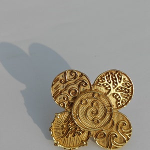 May include: A gold brooch with a four-petal design. Each petal is a circular shape with intricate embossed patterns, including swirls, lines, and a tree-like motif. The brooch is highly reflective, suggesting a polished finish.