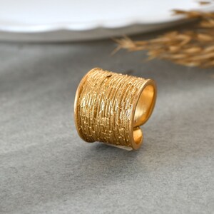 May include: A gold-toned ring with a textured, ridged design. The ring is wide and has a cylindrical shape.