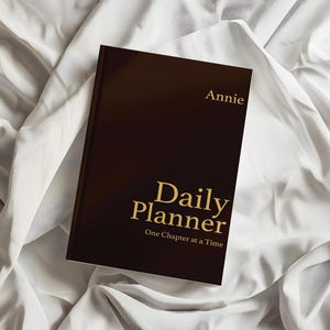 Peut inclure: Un agenda quotidien marron foncé avec des lettres dorées sur la couverture avant. Le texte indique Annie, Daily Planner, et One Chapter at a Time. L'agenda est rectangulaire avec une couverture unie et une typographie claire et lisible.