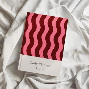 May include: A daily planner with a vertical wavy pattern in alternating shades of dark red and pink on the upper portion. The lower portion is a solid white band featuring the black text Daily Planner and the name Annie centred below it.