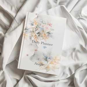 May include: A white daily planner with a watercolor floral design in soft shades of peach, blue, and grey. The cover features the text Daily Planner and the name Annie in a black serif font, centered amidst the delicate botanical illustrations.