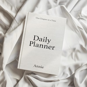 Puede incluir: Una agenda diaria blanca con cubierta texturizada que presenta el texto impreso en negro One Chapter at a Time, Daily Planner y el nombre Annie. La agenda es rectangular con un diseño neutro y una tipografía negra clara y legible.