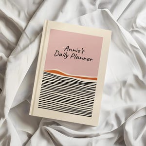 May include: A hardback daily planner with a cream-coloured background. The front cover features a pink rectangular block at the top with the text Annie's Daily Planner in black, followed by a wavy orange line and a series of horizontal black lines.