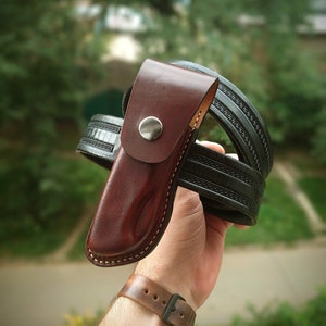 May include: A brown leather knife sheath with a metal snap closure. The sheath is attached to a black leather belt with a decorative pattern.