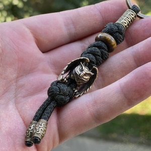 May include: A black paracord keychain with a brass owl charm and a tiger's eye bead. The keychain has a brass ring at the top and two brass beads at the bottom.
