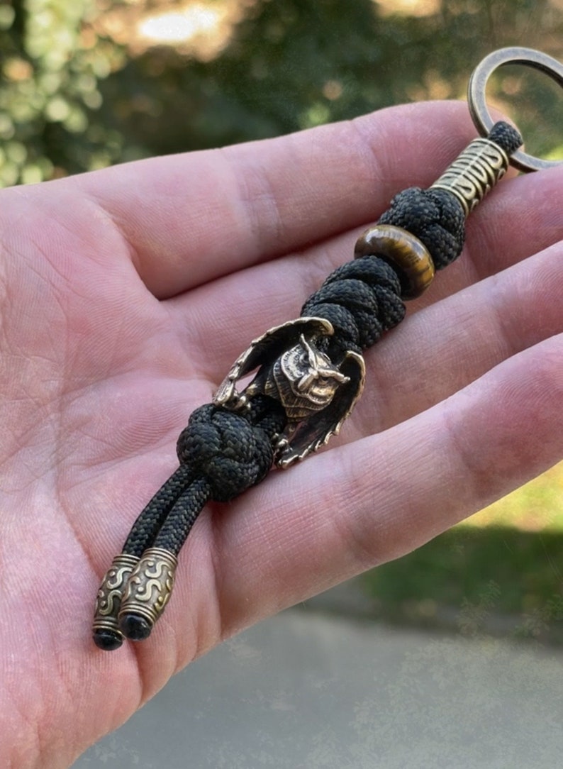 May include: A black paracord keychain with a brass owl charm and a tiger's eye bead. The keychain has a brass ring at the top and two brass beads at the bottom.