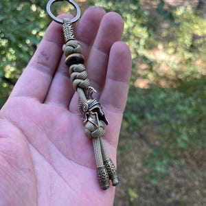 K&ouml;nnte beinhalten: Ein gr&uuml;ner Paracord-Schl&uuml;sselanh&auml;nger mit einem r&ouml;mischen Helm-Anh&auml;nger aus Metall und zwei Metallperlen. Der Schl&uuml;sselanh&auml;nger wird in einer Hand gehalten.