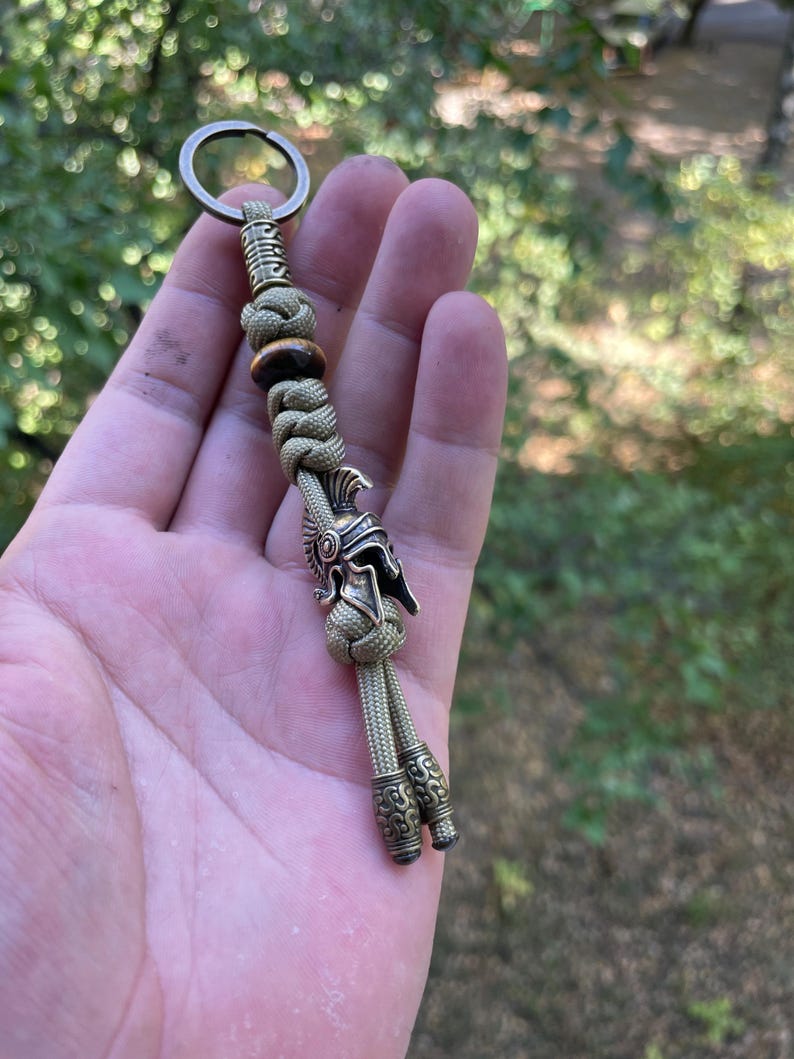 K&ouml;nnte beinhalten: Ein gr&uuml;ner Paracord-Schl&uuml;sselanh&auml;nger mit einem r&ouml;mischen Helm-Anh&auml;nger aus Metall und zwei Metallperlen. Der Schl&uuml;sselanh&auml;nger wird in einer Hand gehalten.