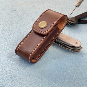 May include: A white ceramic mug with a black handle and the text "But First, Coffee" in a bold, black font. The mug is set against a blurred background, suggesting a cozy setting.