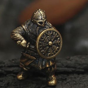 May include: A bronze-colored figurine of a Viking warrior, complete with a helmet, chainmail, and a round shield. The shield features an intricate design. The warrior is posed in a stance, suggesting readiness for battle. The overall aesthetic is detailed and historical.