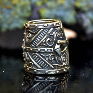 May include: A silver-toned metal bead featuring a detailed design reminiscent of a knight's helmet. The bead has a textured surface with intricate carvings and a raised design on the front.