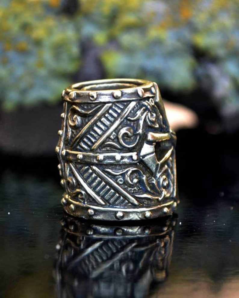 May include: A silver-toned metal bead featuring a detailed design reminiscent of a knight's helmet. The bead has a textured surface with intricate carvings and a raised design on the front.