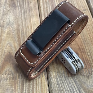 May include: A brown leather pocket knife sheath with a black metal clip and white stitching. A silver and gray pocket knife is partially visible. The sheath is designed to hold a small multi-tool.