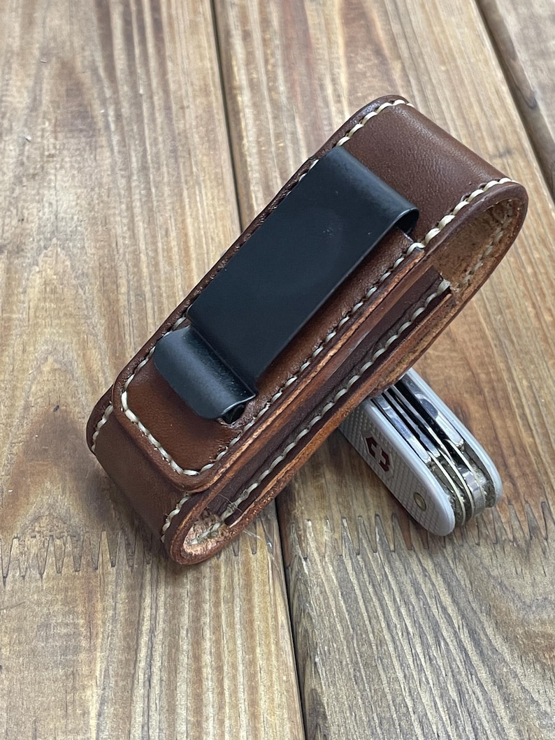 May include: A brown leather pocket knife sheath with a black metal clip and white stitching. A silver and gray pocket knife is partially visible. The sheath is designed to hold a small multi-tool.