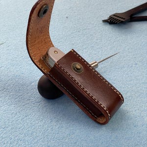 May include: A brown leather sheath with a silver multi-tool and a metal awl. The sheath is secured with a snap closure and features white stitching. The awl is partially inserted into the sheath. The background is a light blue surface.