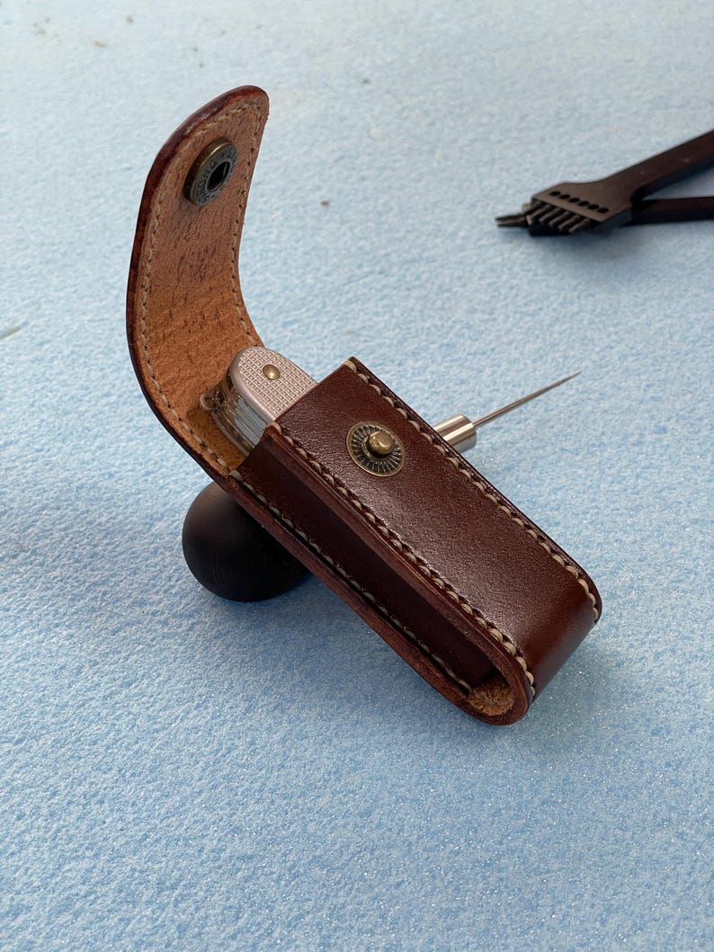 May include: A brown leather sheath with a silver multi-tool and a metal awl. The sheath is secured with a snap closure and features white stitching. The awl is partially inserted into the sheath. The background is a light blue surface.