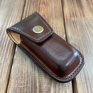 Op de afbeelding: Een bruin leren etui met een messing knoopsluiting. De etui is ontworpen om een ​​zakmes of ander klein gereedschap te bevatten.