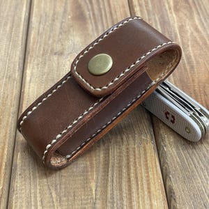 May include: A brown leather knife sheath with white stitching and a brass button closure. A silver and white pocket knife is partially visible next to the sheath. The items are on a wooden surface.